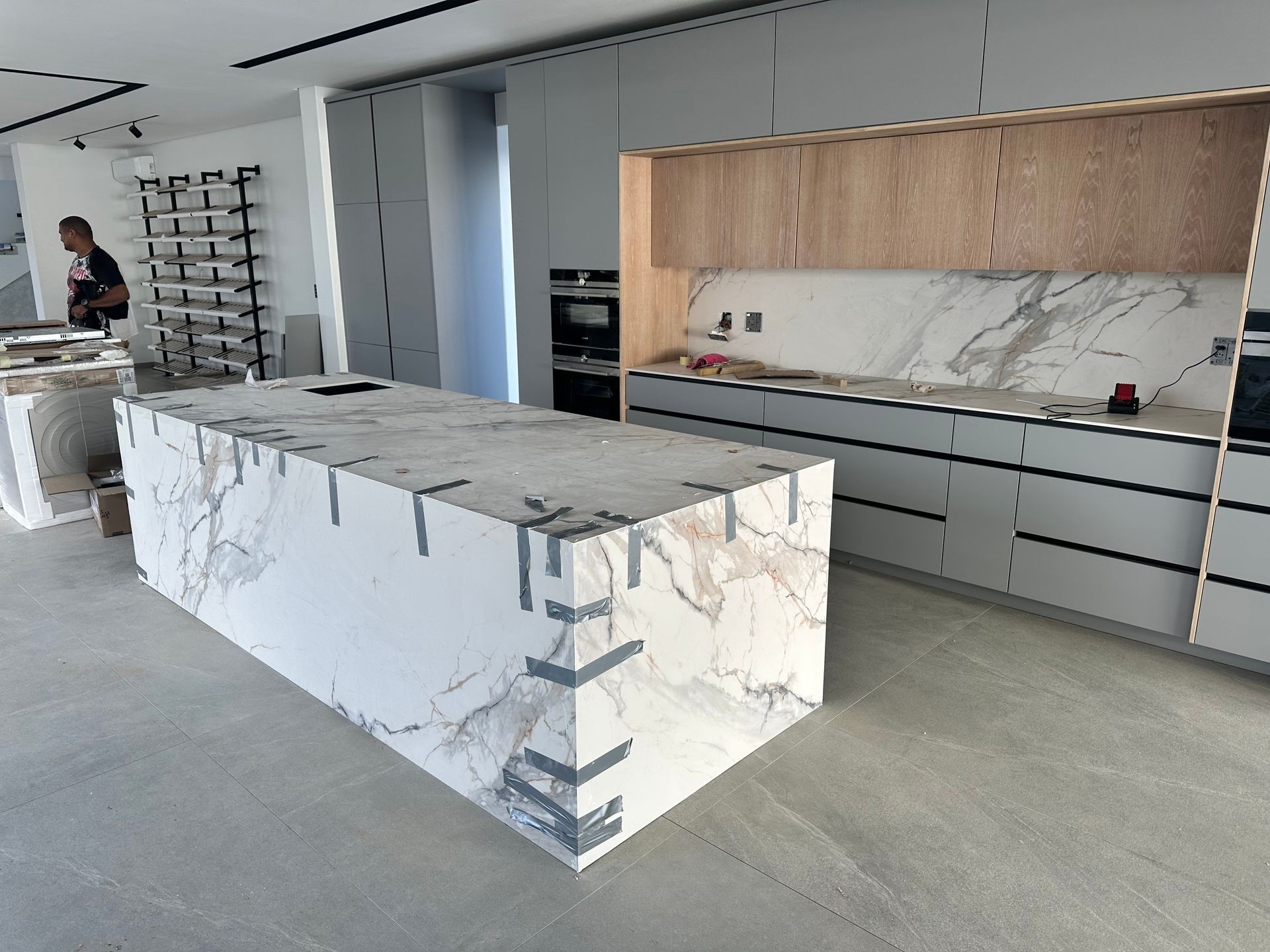Modern kitchen under construction with marble island and wooden cabinets, showing a person working in the background.